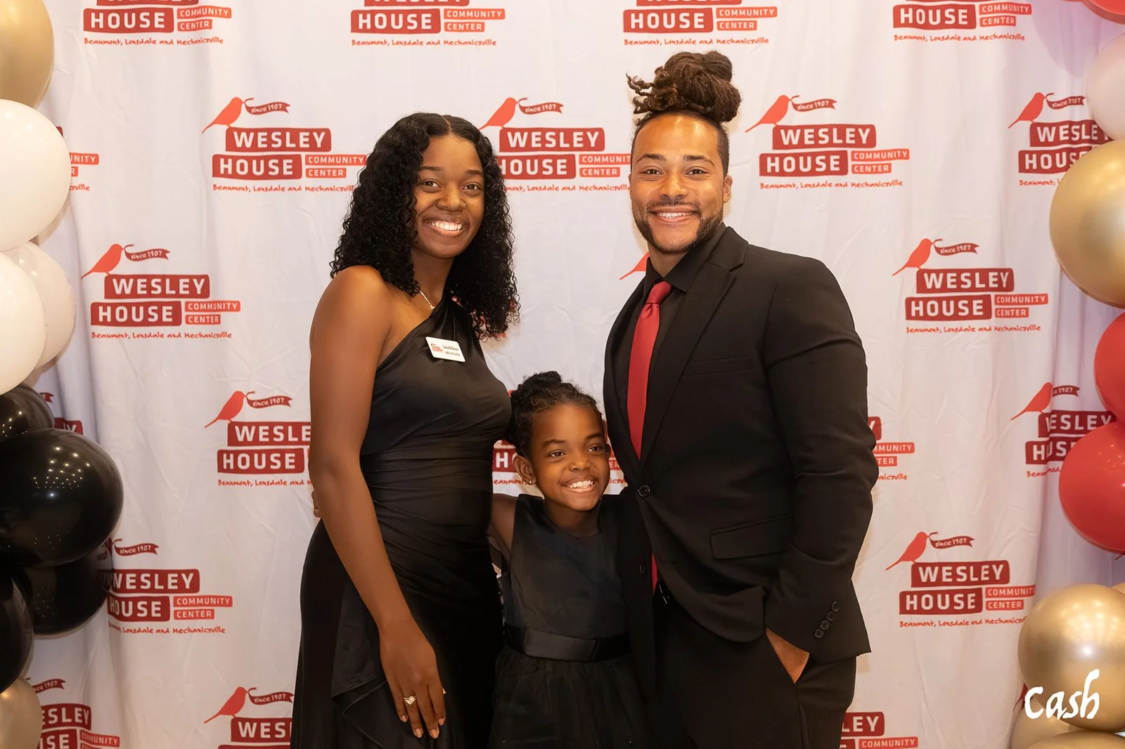 A family of three standing together at an event with a Wesley House Community Center backdrop. The woman is wearing a black dress, the man is in a dark suit with a red tie, and the girl is dressed in black, smiling with their arms around each other.