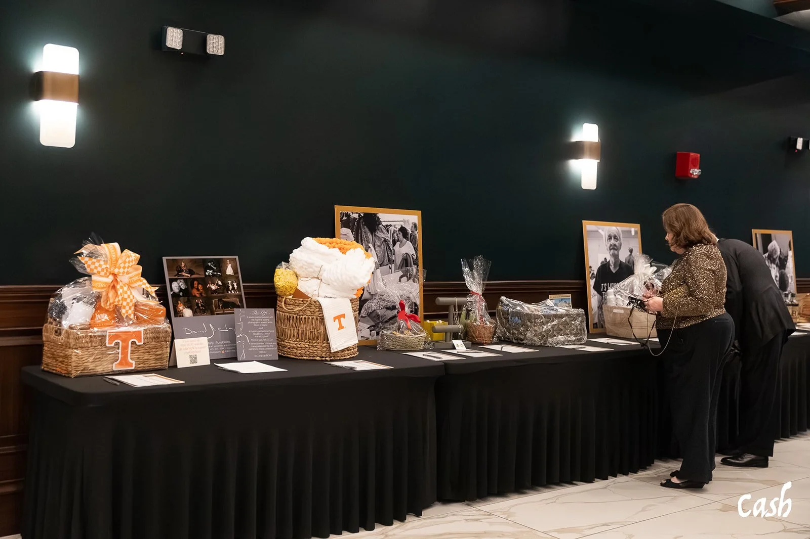 A long table with various gift baskets and framed photographs, people looking at the items, dark wall with wall lights in the background.