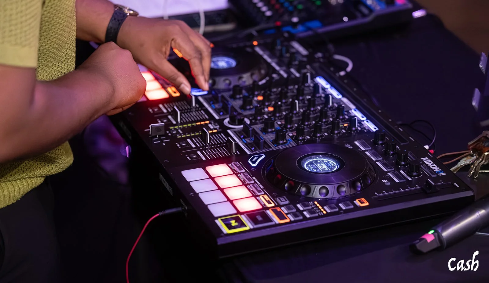 Close-up of a DJ's hands operating a professional DJ mixer and controller with vibrant, illuminated buttons and knobs, in a dimly lit setting.