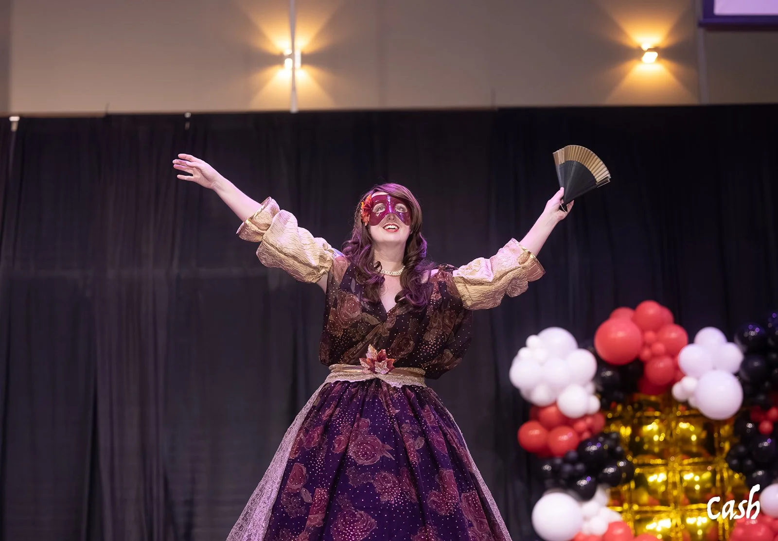 Woman in a fancy dress and mask on stage with balloons in the background