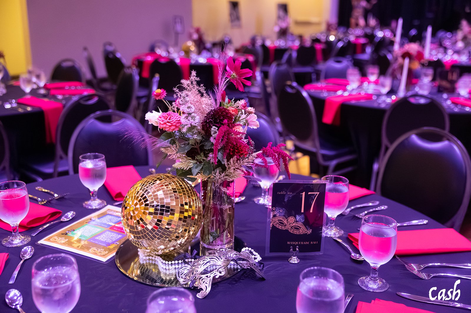 A banquet table decorated with pink flowers, a gold disco ball, and pink napkins at a Masquerade Ball event.
