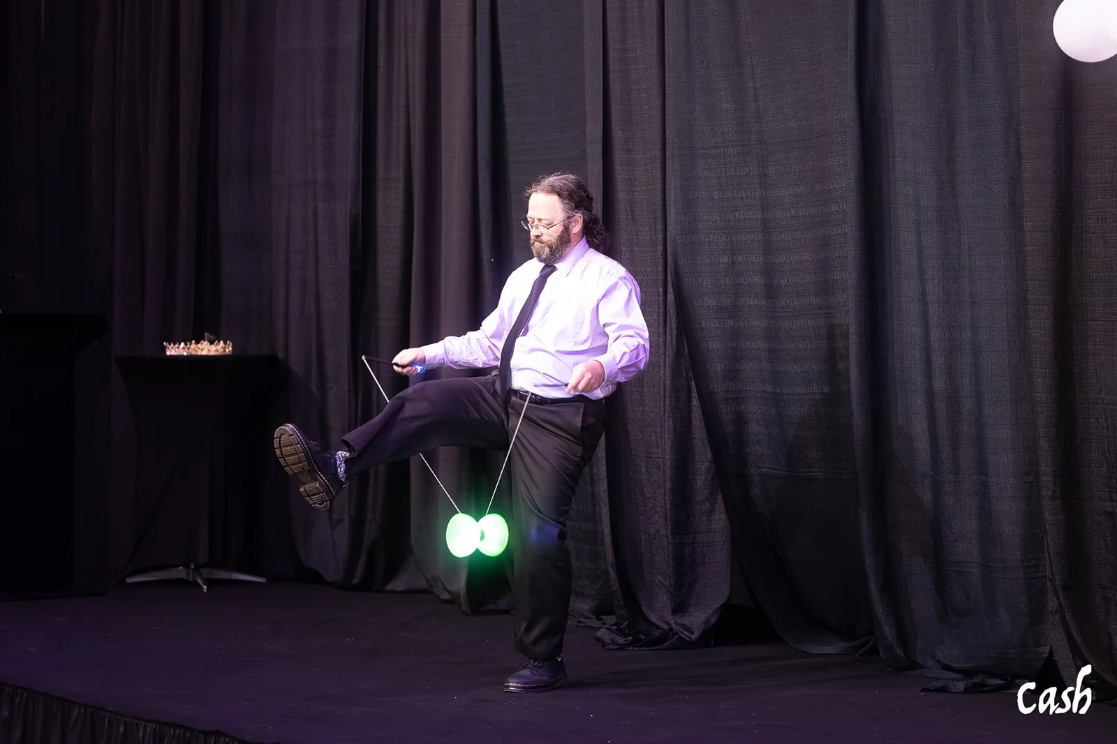 A man in a white shirt, black tie, and black pants on a stage with black curtains in the background. He appears to be performing a yo-yo trick, with a glowing green yo-yo caught on his foot and strings in his hands. The word 'Cash' is written in whit