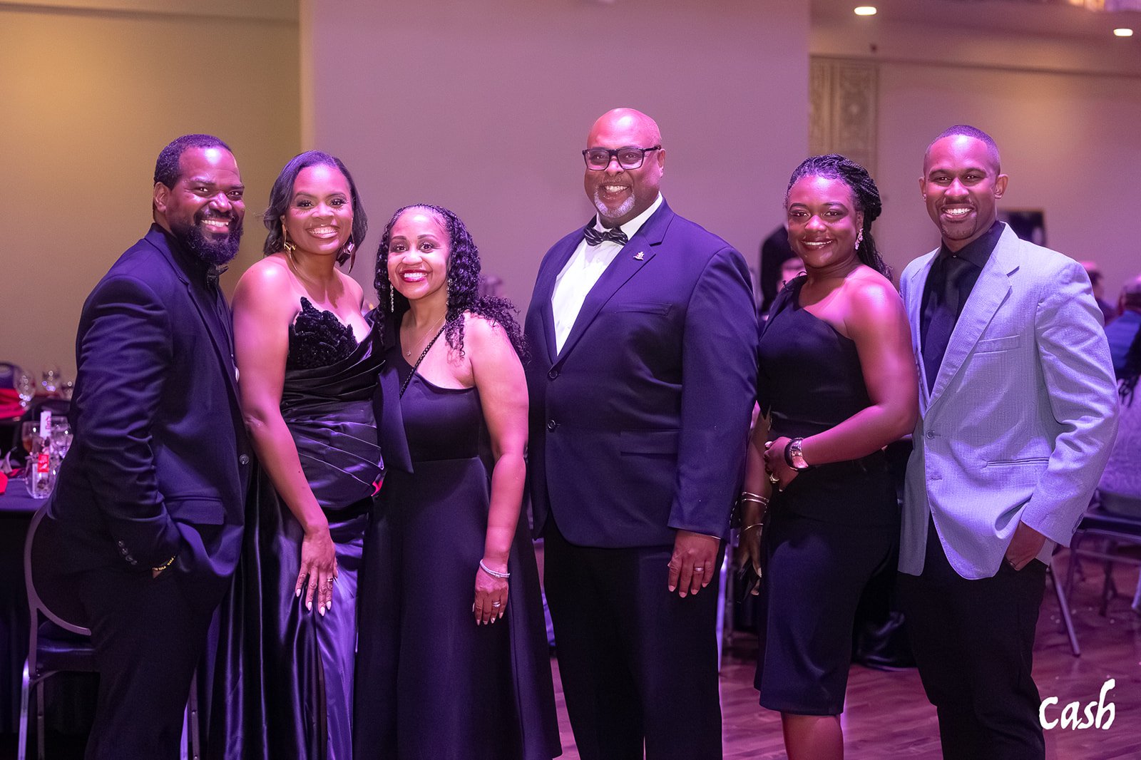 Seven people dressed in formal attire standing together at an indoor event, smiling at the camera.