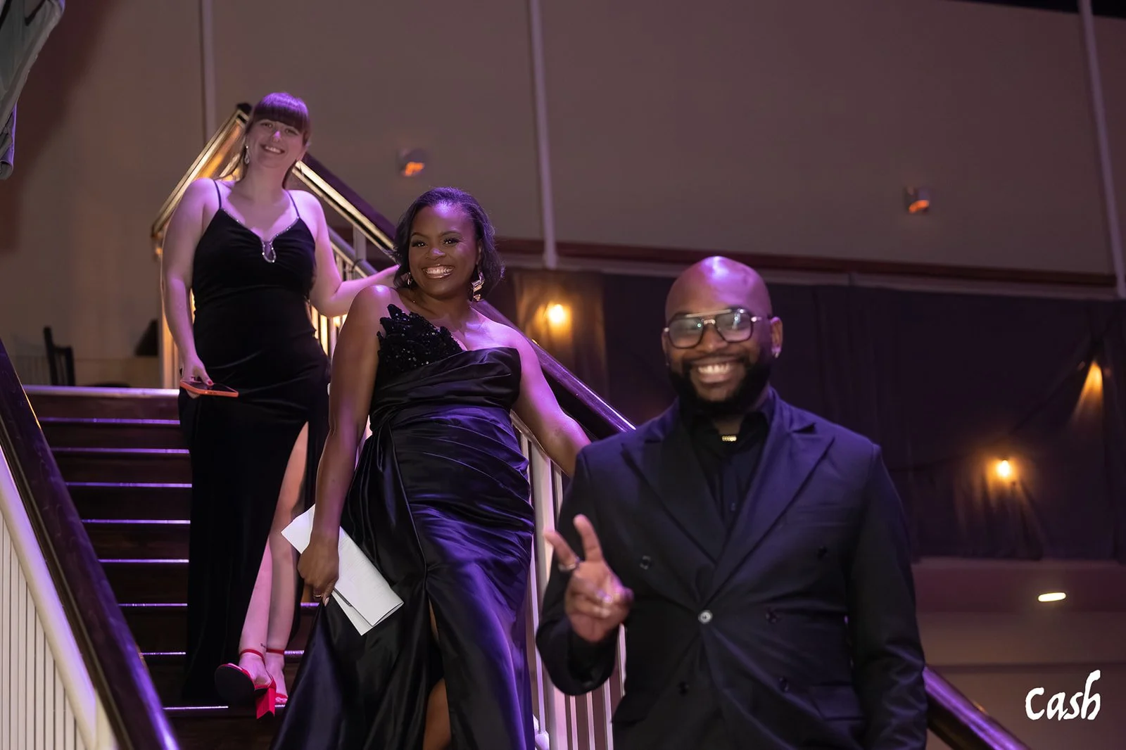 Three people dressed in formal black attire standing on a staircase at an event. The goal is smiling, with a woman in a black gown with a slit, a woman in a black dress with a slit, and a man in a black suit with glasses.