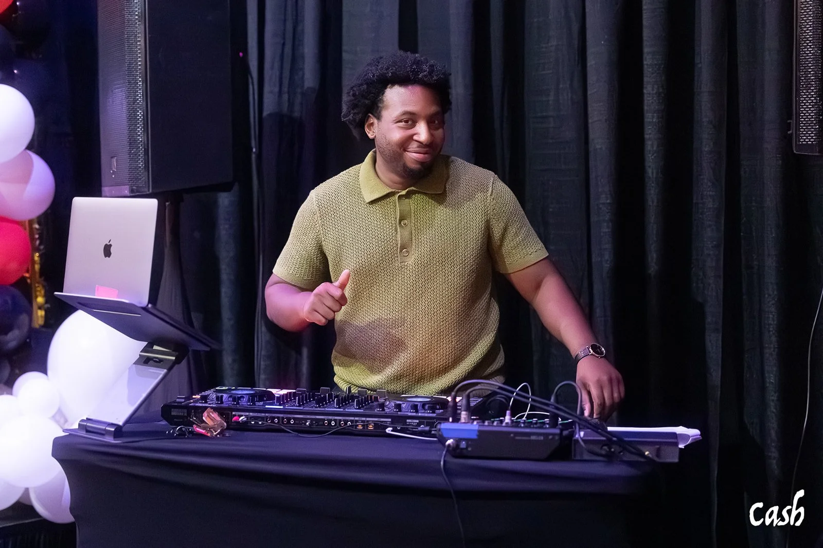 A man with curly hair and a beard, wearing a yellow-green polo shirt, is DJing at a party with a black DJ controller on a table. There are balloons on the left and a black curtain in the background.