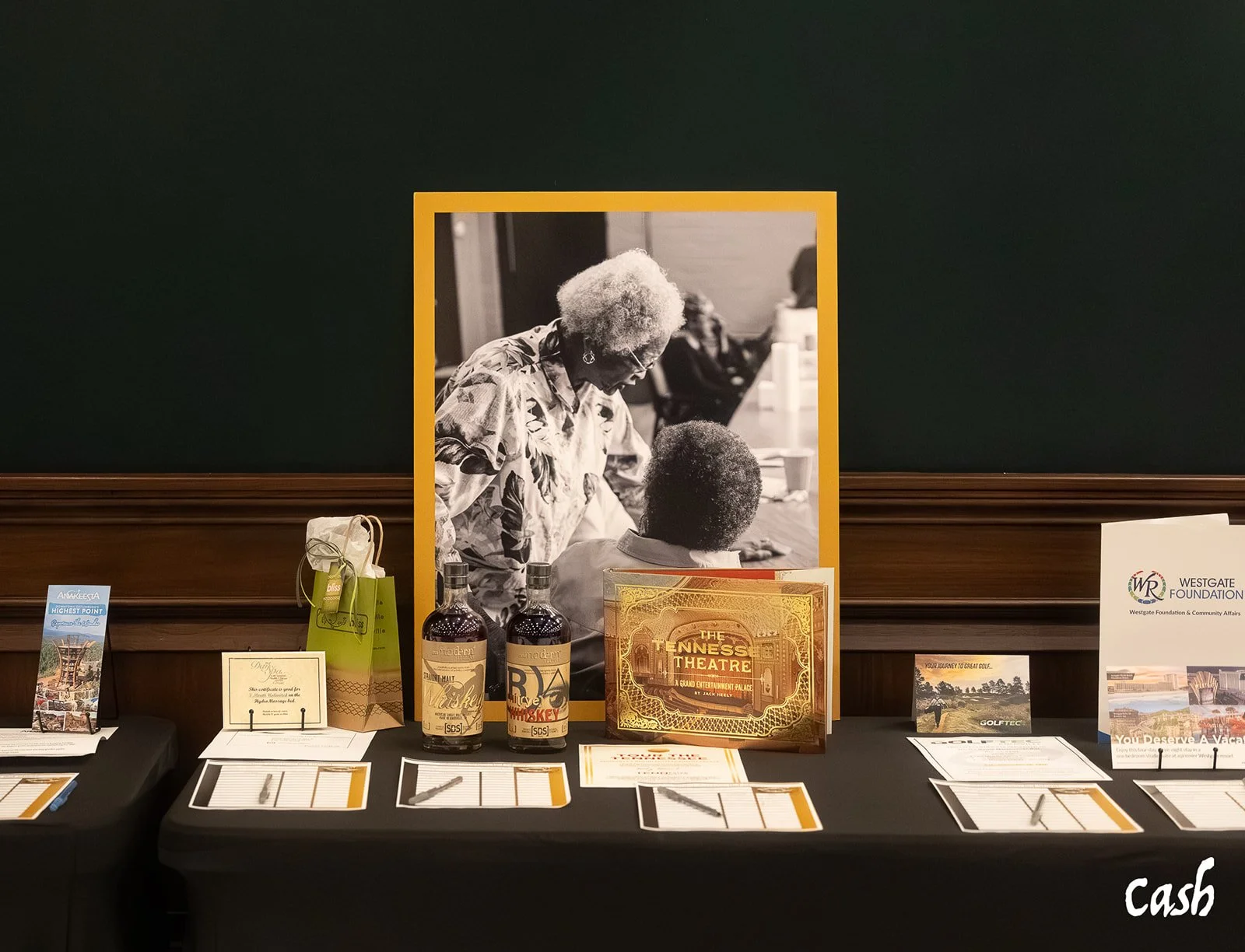 A display table with framed photos, bottles of alcohol, brochures, and registration forms, with a blackboard in the background.