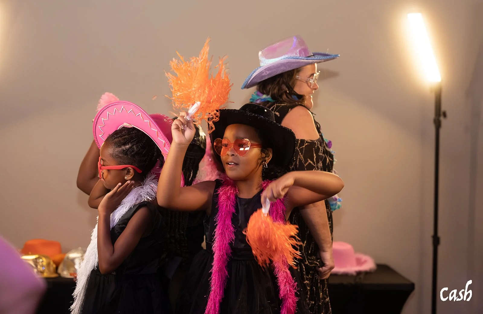 Two young girls and an adult woman dressed in colorful costumes with hats, boas, and sunglasses, posing in a room with pink and orange accessories in the background.