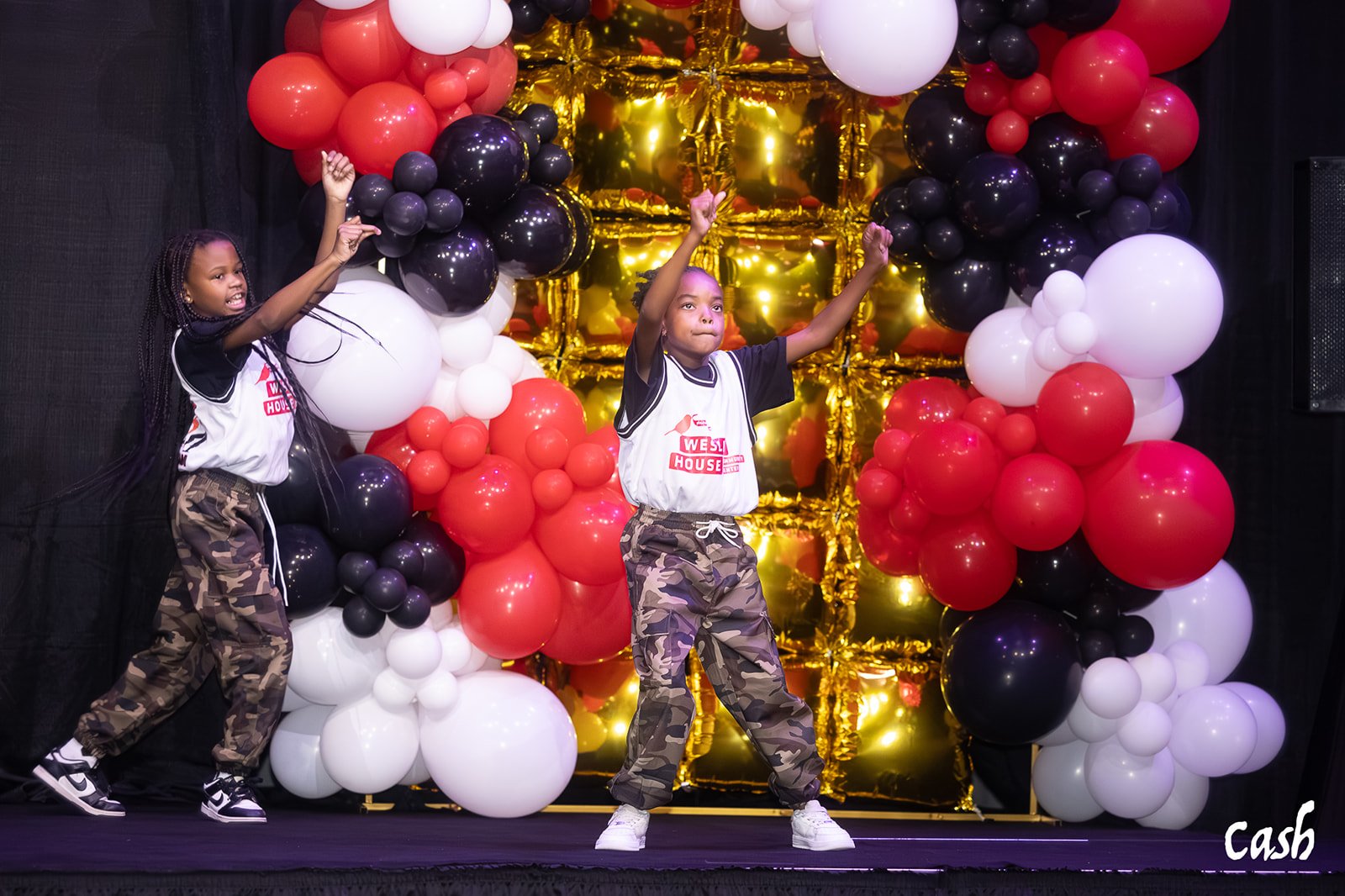 Two young girls dancing on a stage decorated with balloons in red, white, black, and gold, with one girl striking a pose and the other raising her arm and smiling.