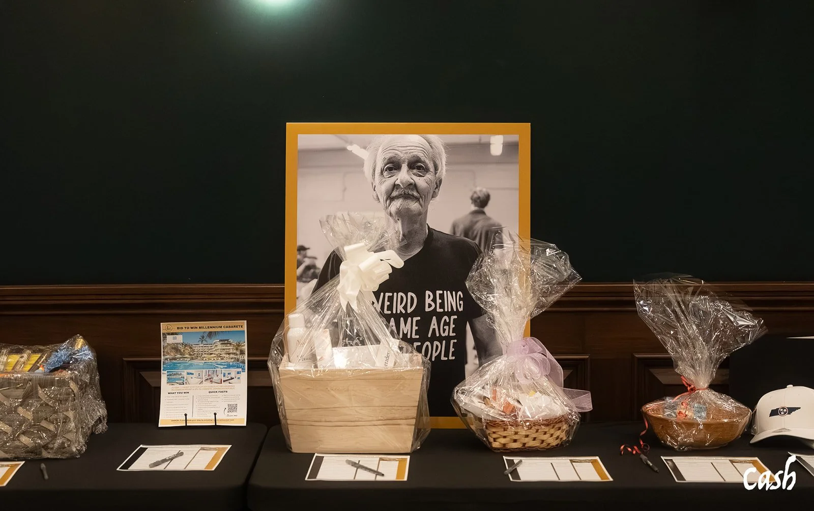 Auction table with prizes, including wrapped gifts and a white helmet, in front of a large black-and-white photo of an elderly woman wearing a black T-shirt with white text, in an indoor setting.