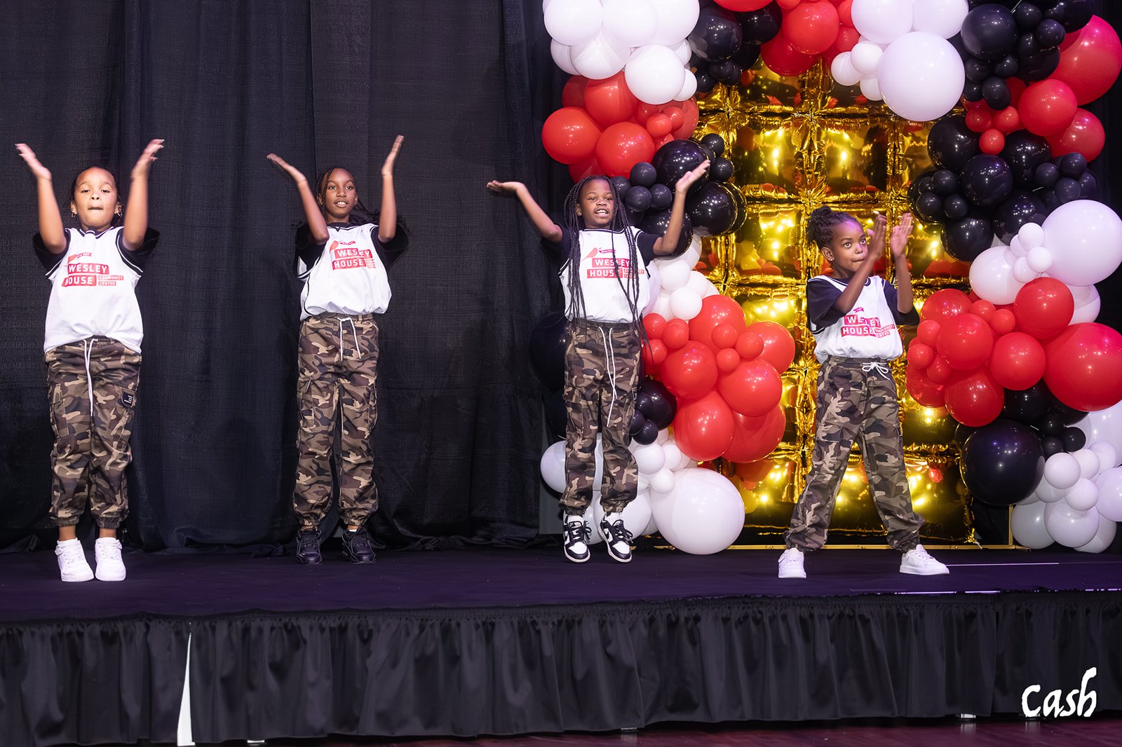 Four young girls perform a dance on stage, wearing camouflage pants and white shirts with red logos. A large balloon display with black, white, red, and gold balloons decorates the backdrop.