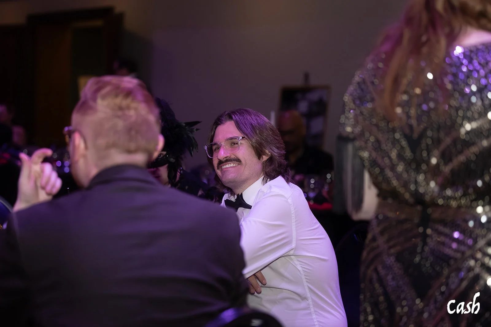 A man with glasses and a mustache, wearing a white shirt and black bow tie, smiling at a social event.