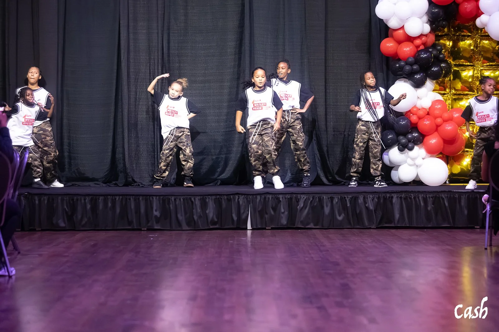 Young girls performing a dance on stage with black curtains, camouflage pants, and casual white sneakers. Decorations include a large cluster of red, black, white, and gold balloons on the right side. Some girls are mid-movement, with one girl in the