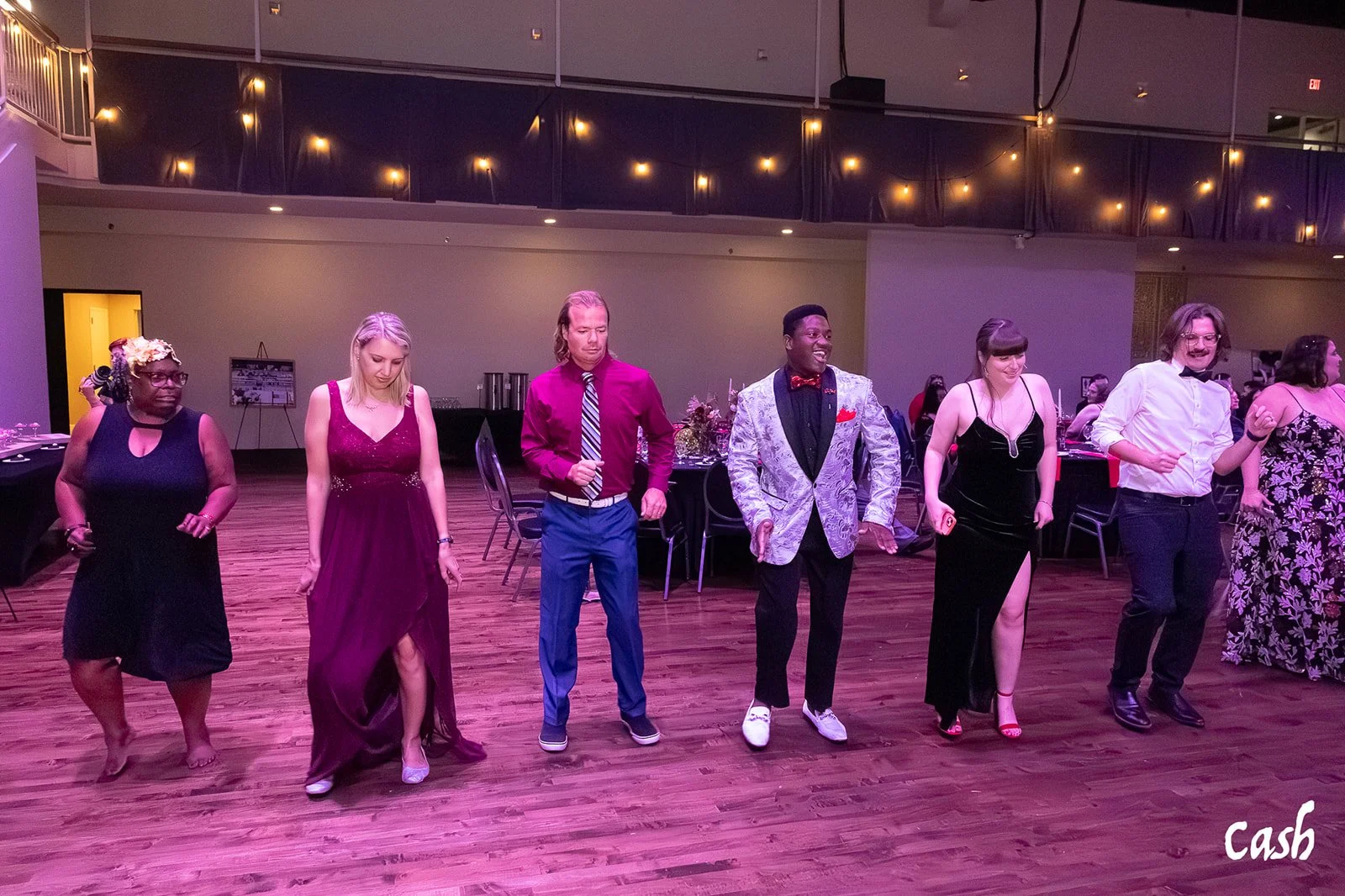 People dancing at a social event in a hall with wood floors and string lights overhead.