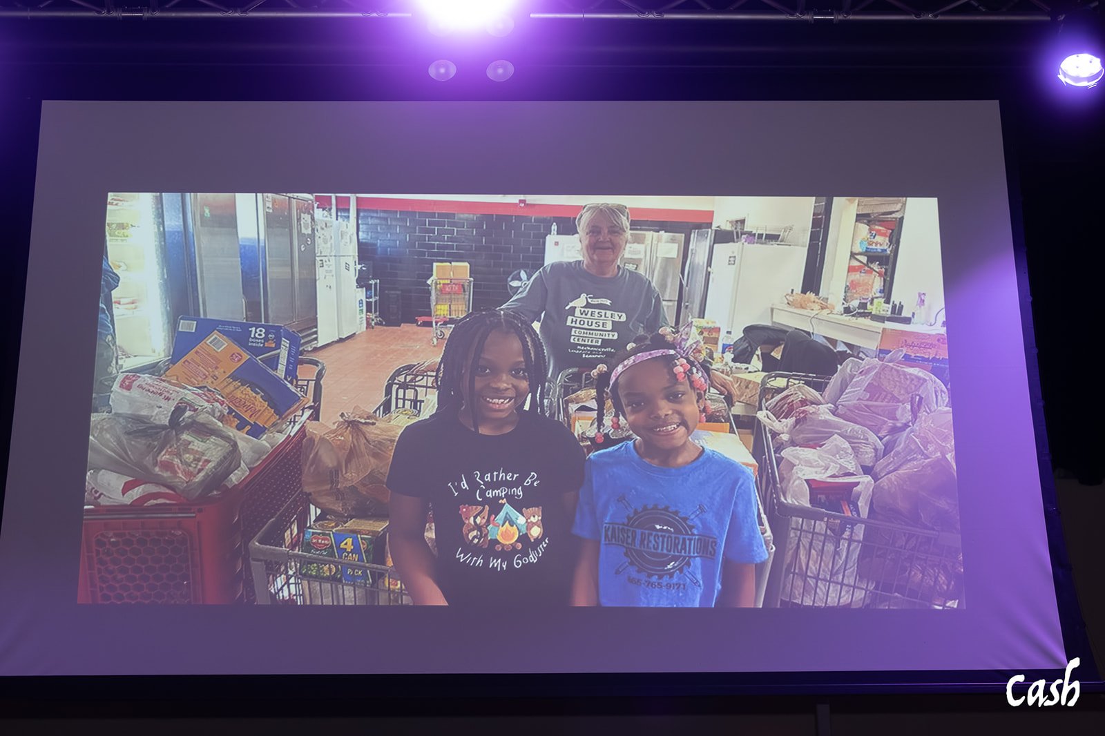 Two young girls smiling in front of a donation area with food and supplies, with an older woman standing behind them, inside a community center.