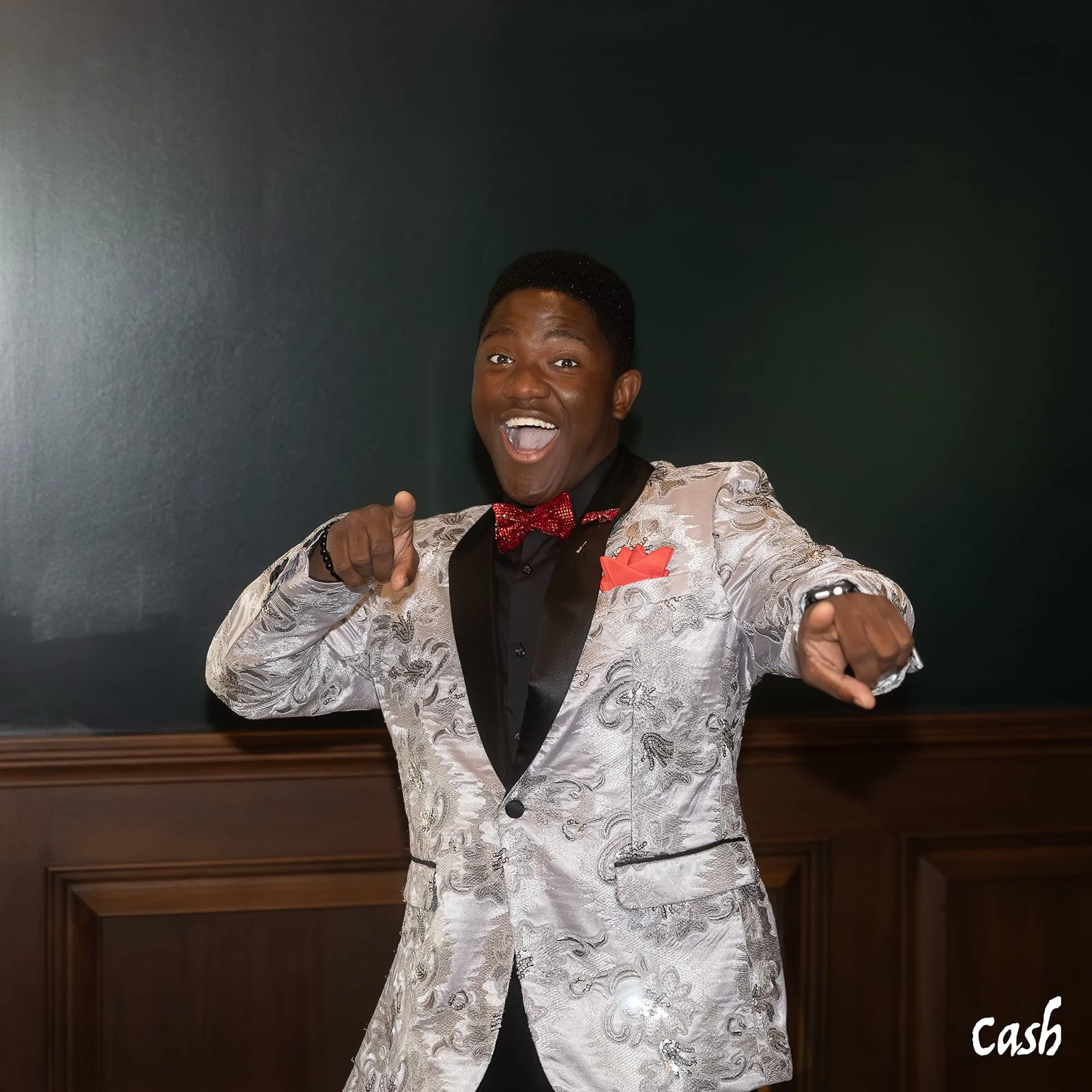 Young man in a shiny silver patterned tuxedo with black lapels, red bow tie, and red pocket square pointing and smiling excitedly.