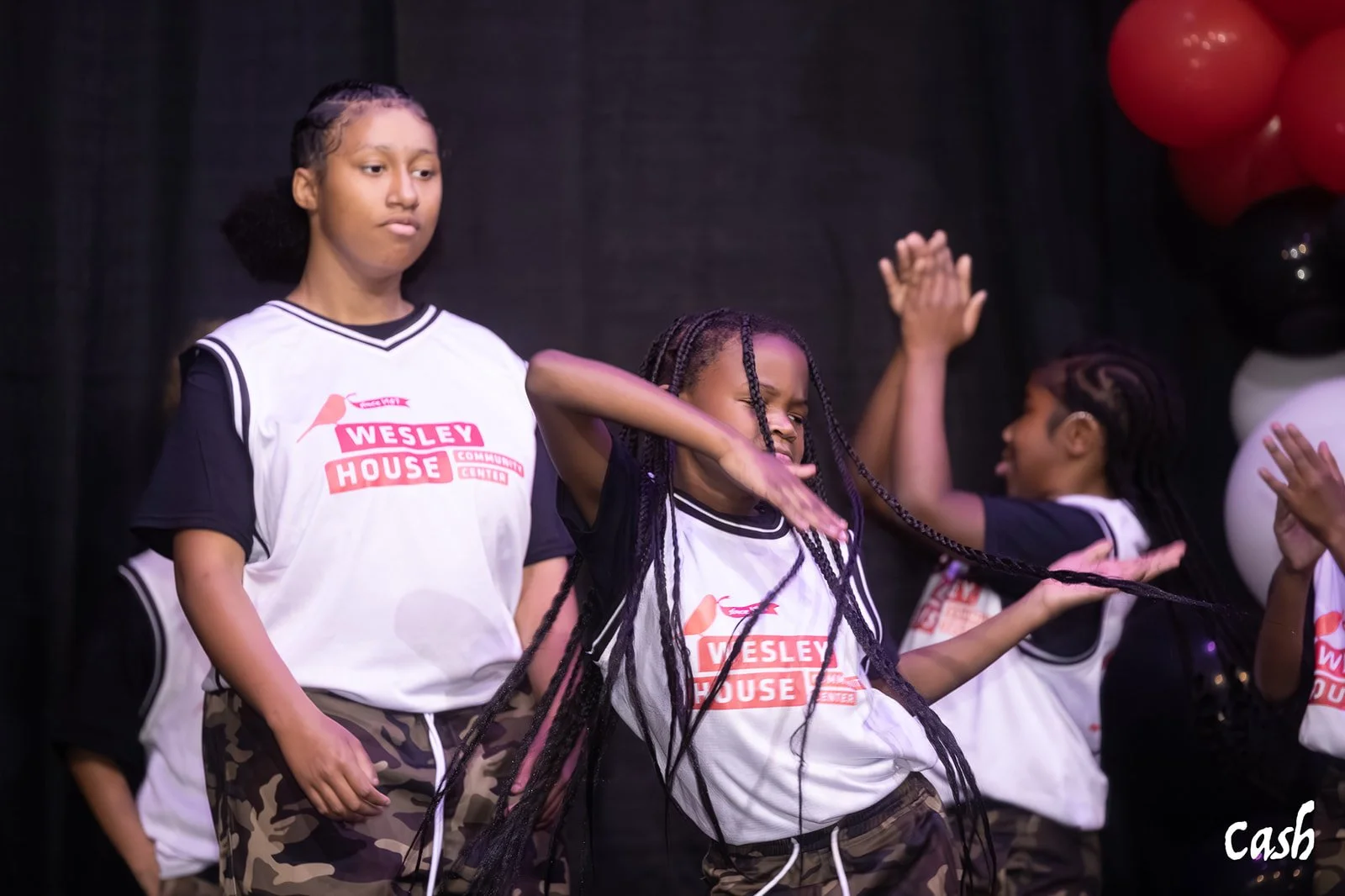 Group of young girls wearing Wesely House community center T-shirts, some dancing on stage with balloons in the background.