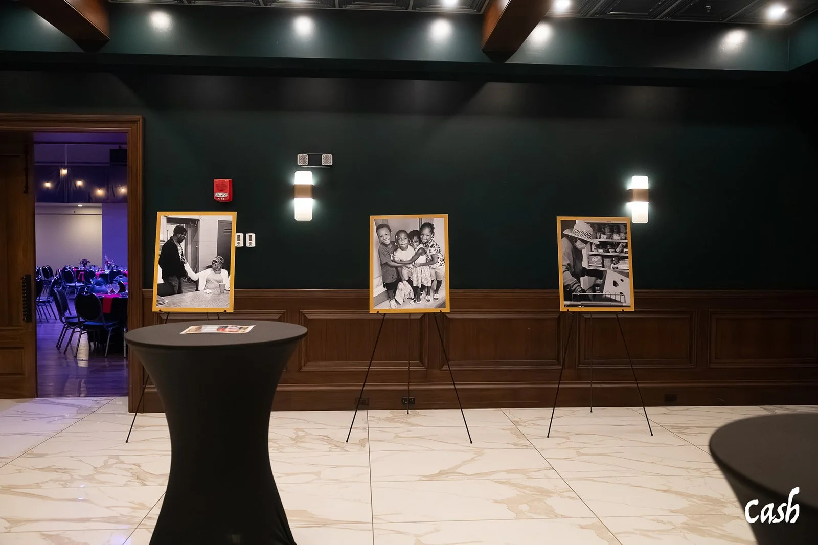 Black-and-white photographs displayed on stands in a gallery or event space, with a dark green wall and wooden paneling, and tables with black tablecloths in the foreground.