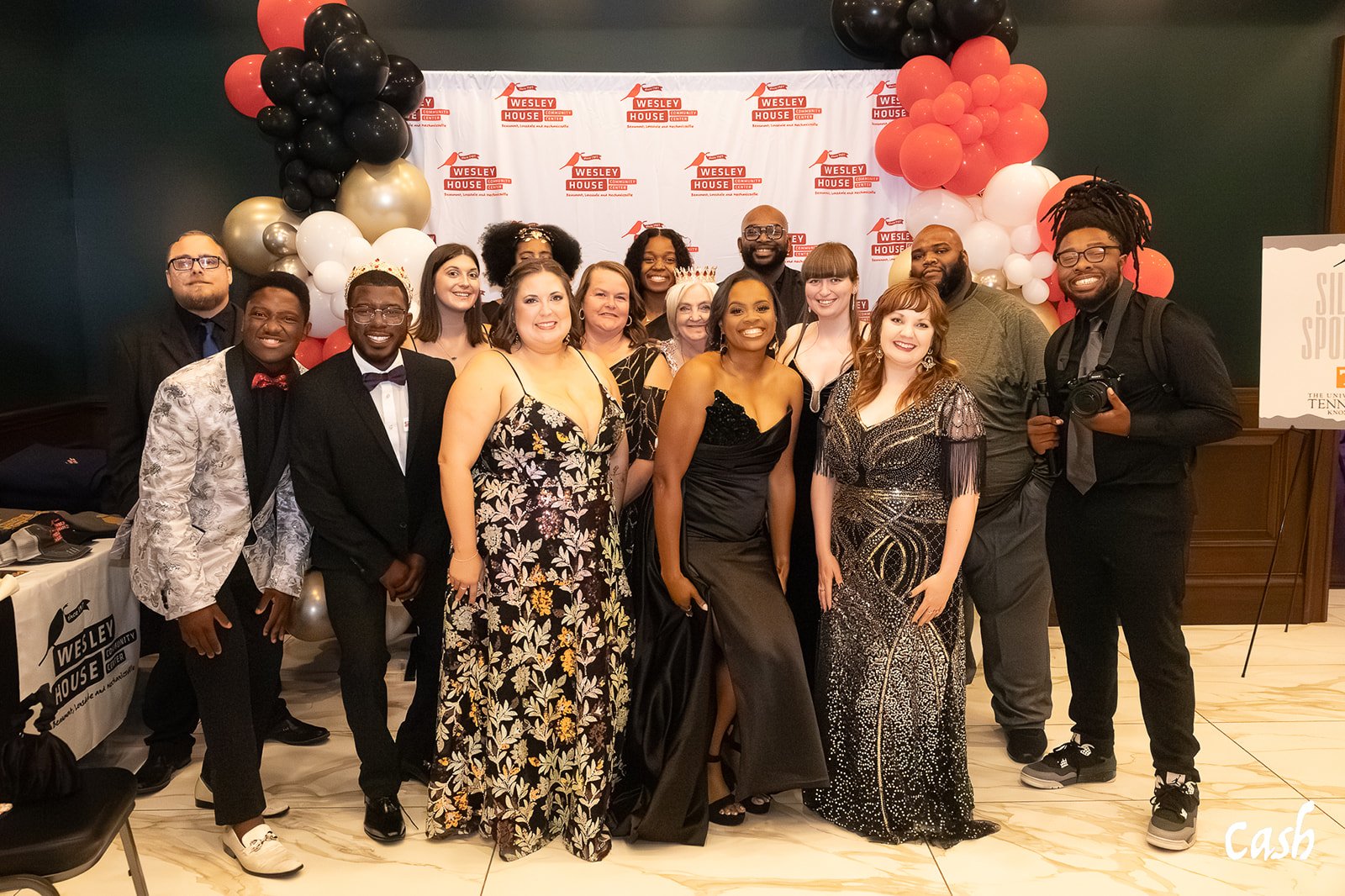 Group of people dressed in formal attire at a celebration event with balloon decorations and a backdrop that reads 'Wesley House.'