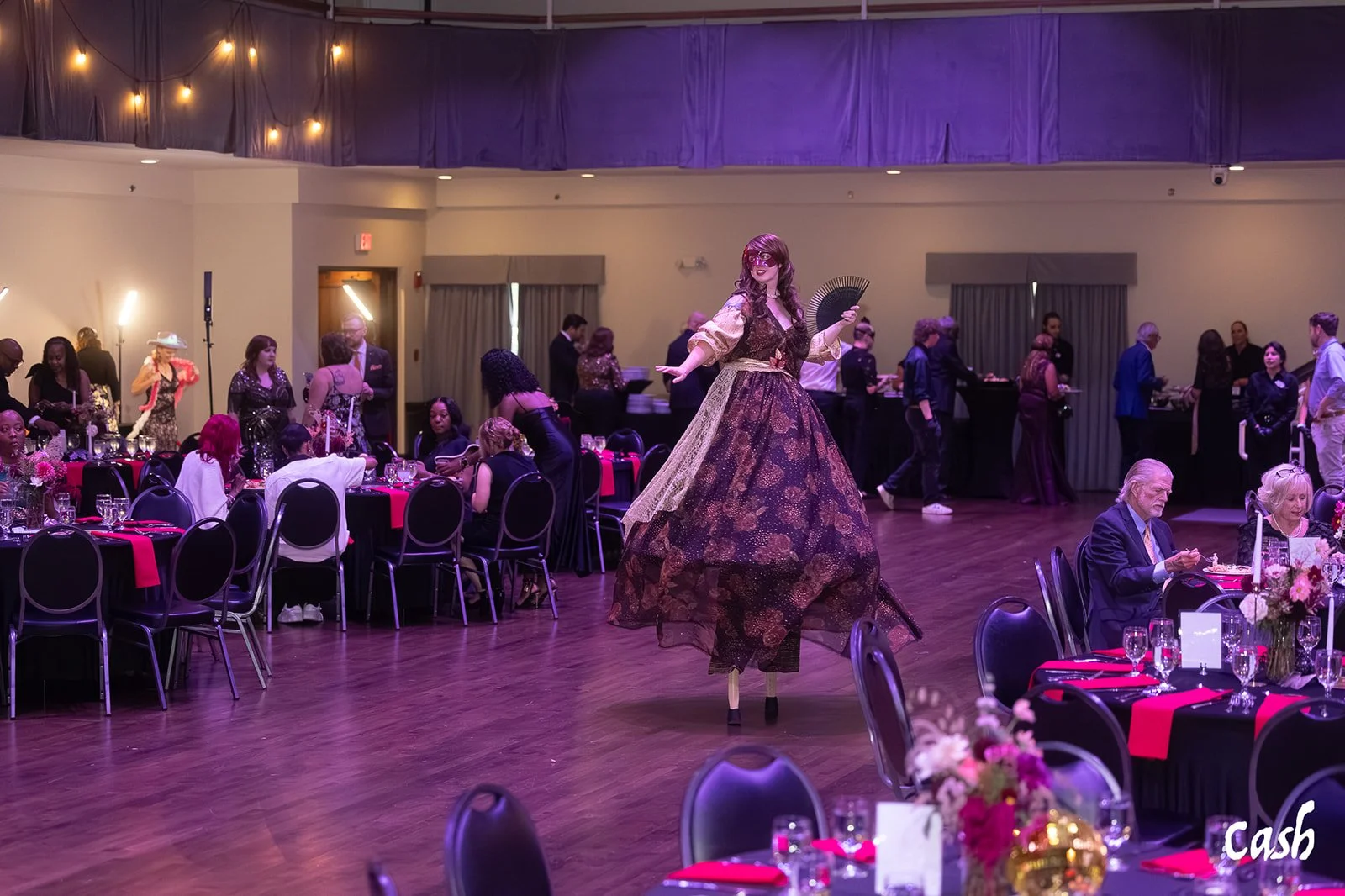 Woman on stilts performing at a decorated event space with round tables, guests, and purple lighting.