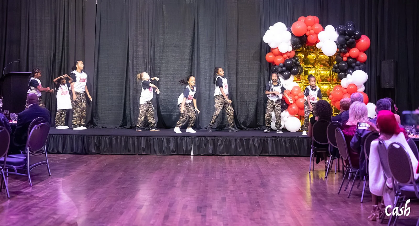 Young girls dressed in camo pants and black and white tops perform a dance on stage decorated with red, black, and white balloons and a gold backdrop. Audience members are seated at tables watching the performance.