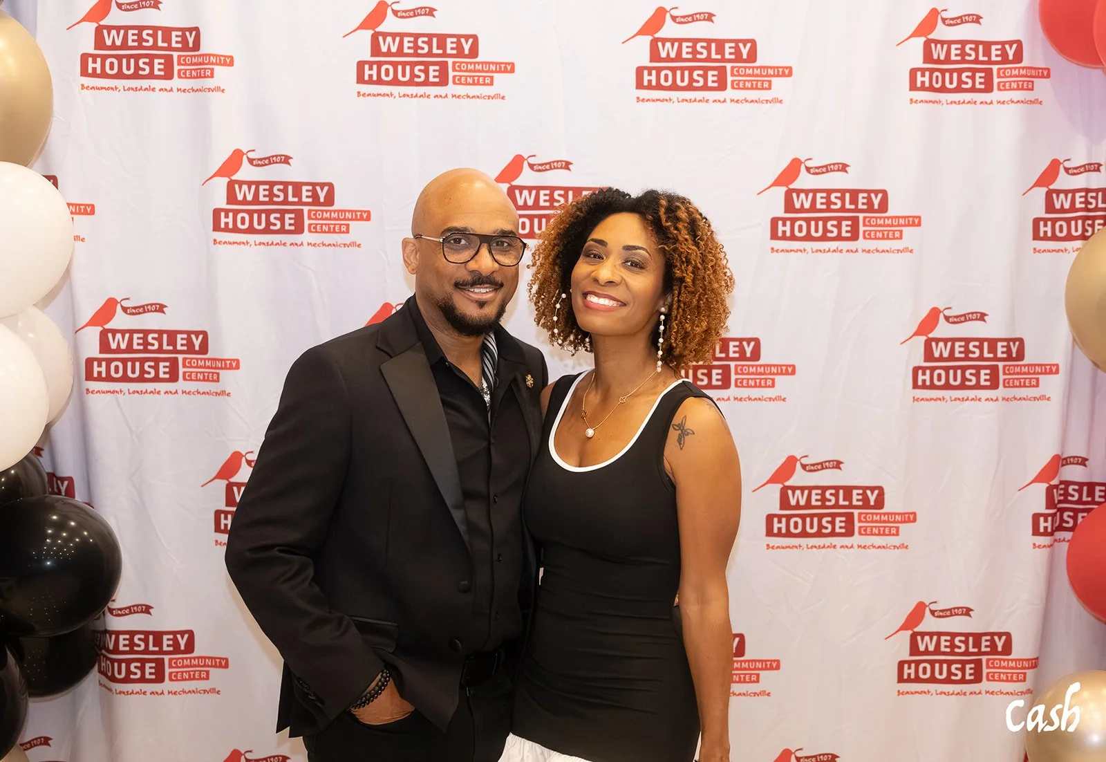 A man and a woman standing close together, smiling, during a event at Wesley House Community Center with a white backdrop featuring the organization's logo and name. The man wears a black suit with a bowtie, and the woman wears a black dress with whi