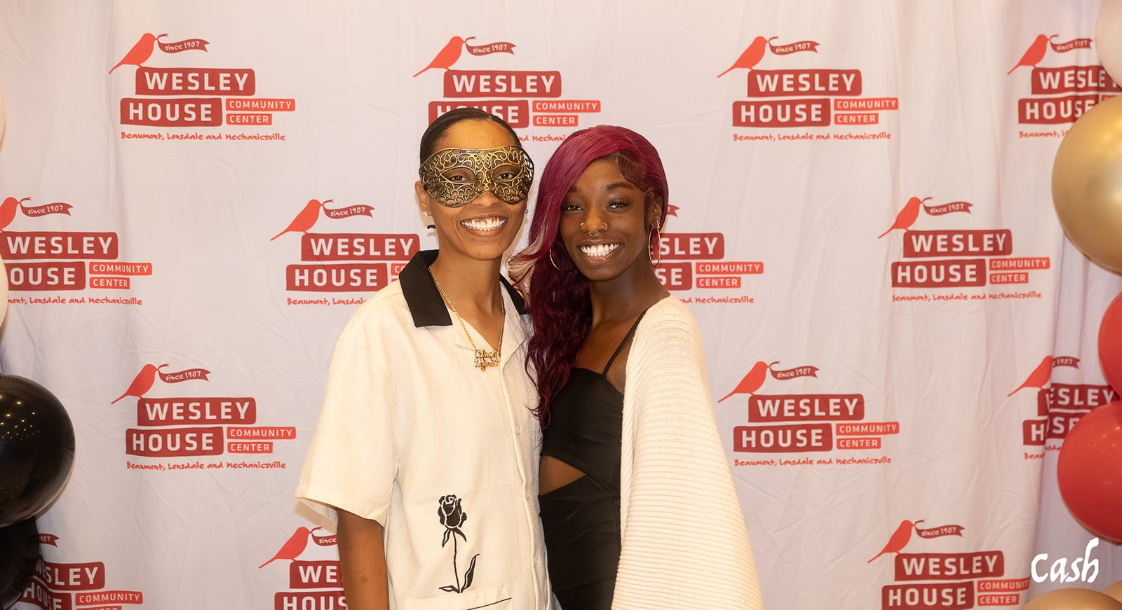 Two women standing in front of a step and repeat banner for Wesley House Community Center. One woman is wearing a gold mask and a light-colored shirt with a black collar, while the other has long, wavy, burgundy hair and is dressed in a black outfit 