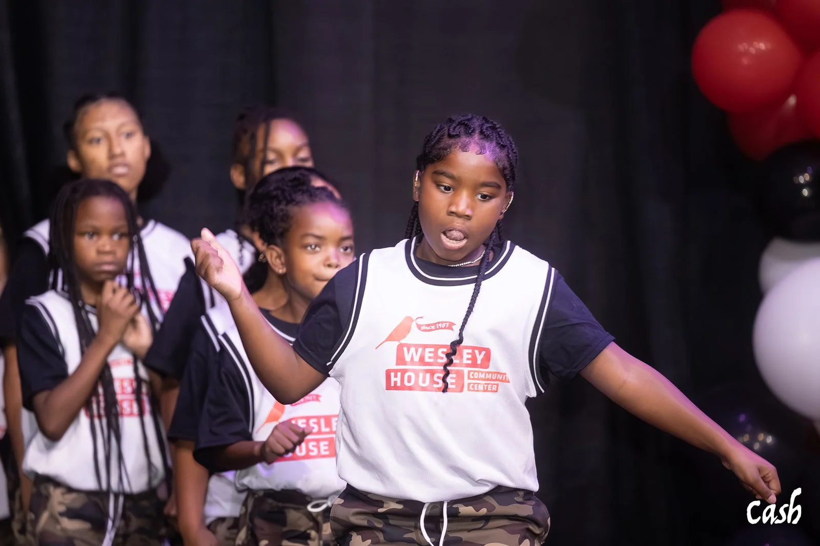 Group of young girls performing on stage, wearing black and white jerseys with 'Wesley House' written on them, with balloons in the background.