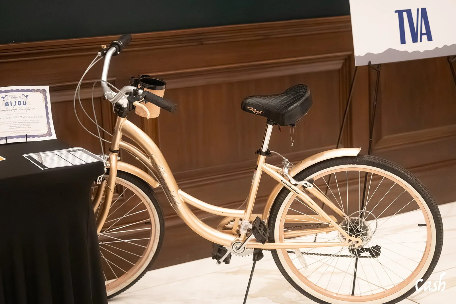 A vintage beige bicycle with black seat and tires, parked indoors near a wooden wall, partially visible signs, and a blurrily positioned black table.