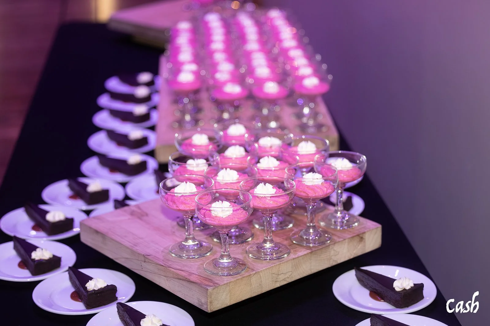 A dessert table with pink and purple treats, including glasses of pink mousse topped with white whipped cream and plates of chocolate cake slices with a dollop of cream.