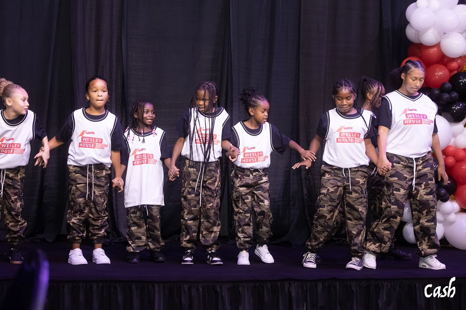 Eight young girls standing on a stage holding hands, dressed in white and black shirts with 'Wesley House' logos, and camo pants, participating in a performance or dance.