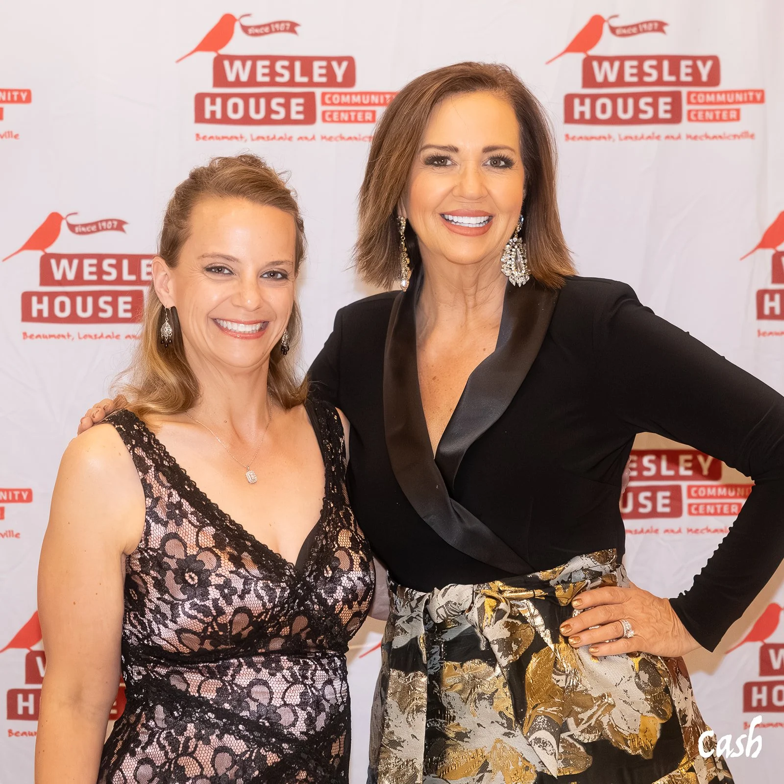 Two women smiling at an event, standing in front of a backdrop with 'Wesley House Community Center' logos, with one woman wearing a black and floral dress, and the other in a black jacket and patterned skirt.