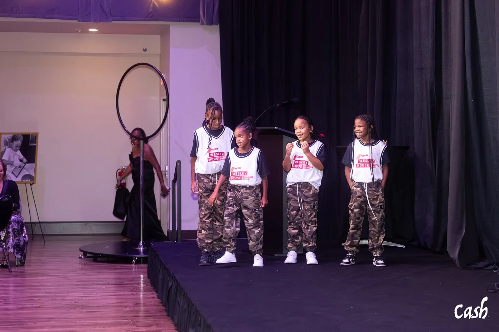 Four young girls wearing white and navy tops with camouflage pants standing on a stage, smiling and talking, with a black curtain behind them. A woman in a black dress walks by in the background, and a fire extinguisher is visible on the wall.