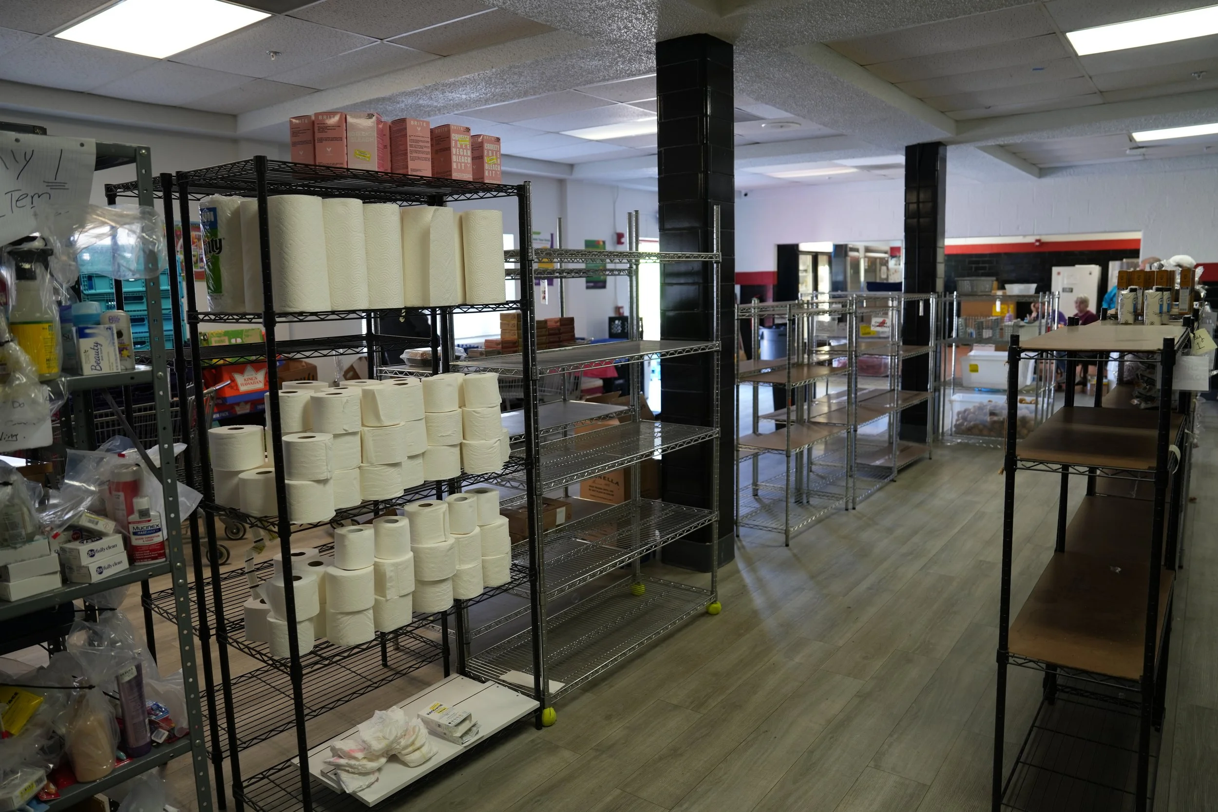 Empty metal shelving units in a store or storage area with some paper towels, supplies, and miscellaneous items, and a few people visible in the background near the checkout area.