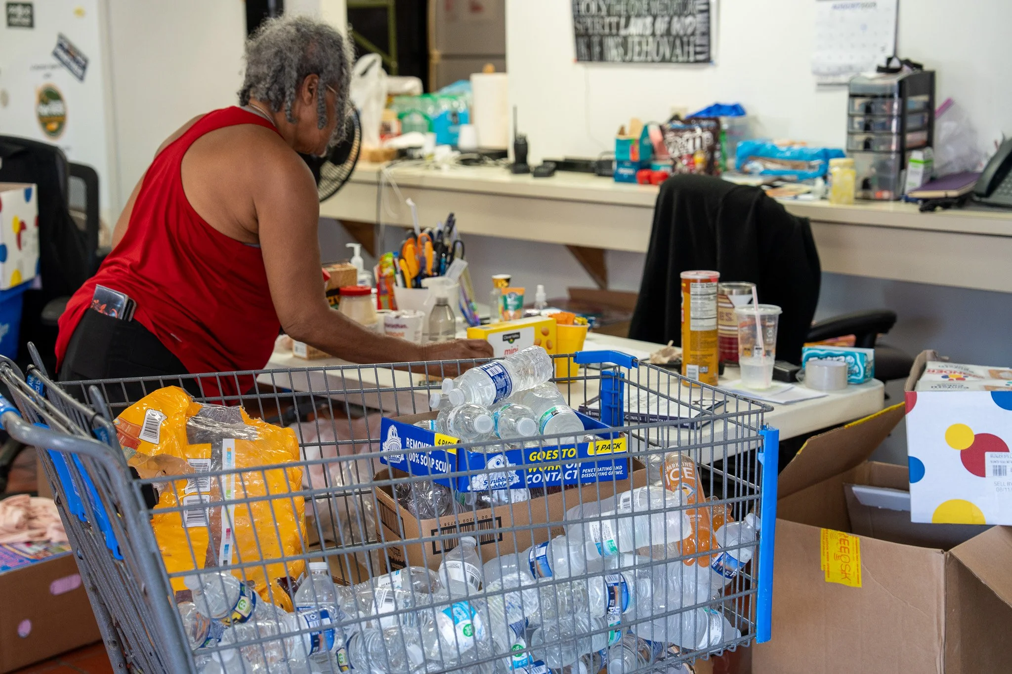A woman with gray, curly hair, wearing a red sleeveless top, stands at a cluttered table sorting through items. The table is covered with various supplies, including plastic bottles, boxes, and papers. In the foreground, there is a shopping cart fill