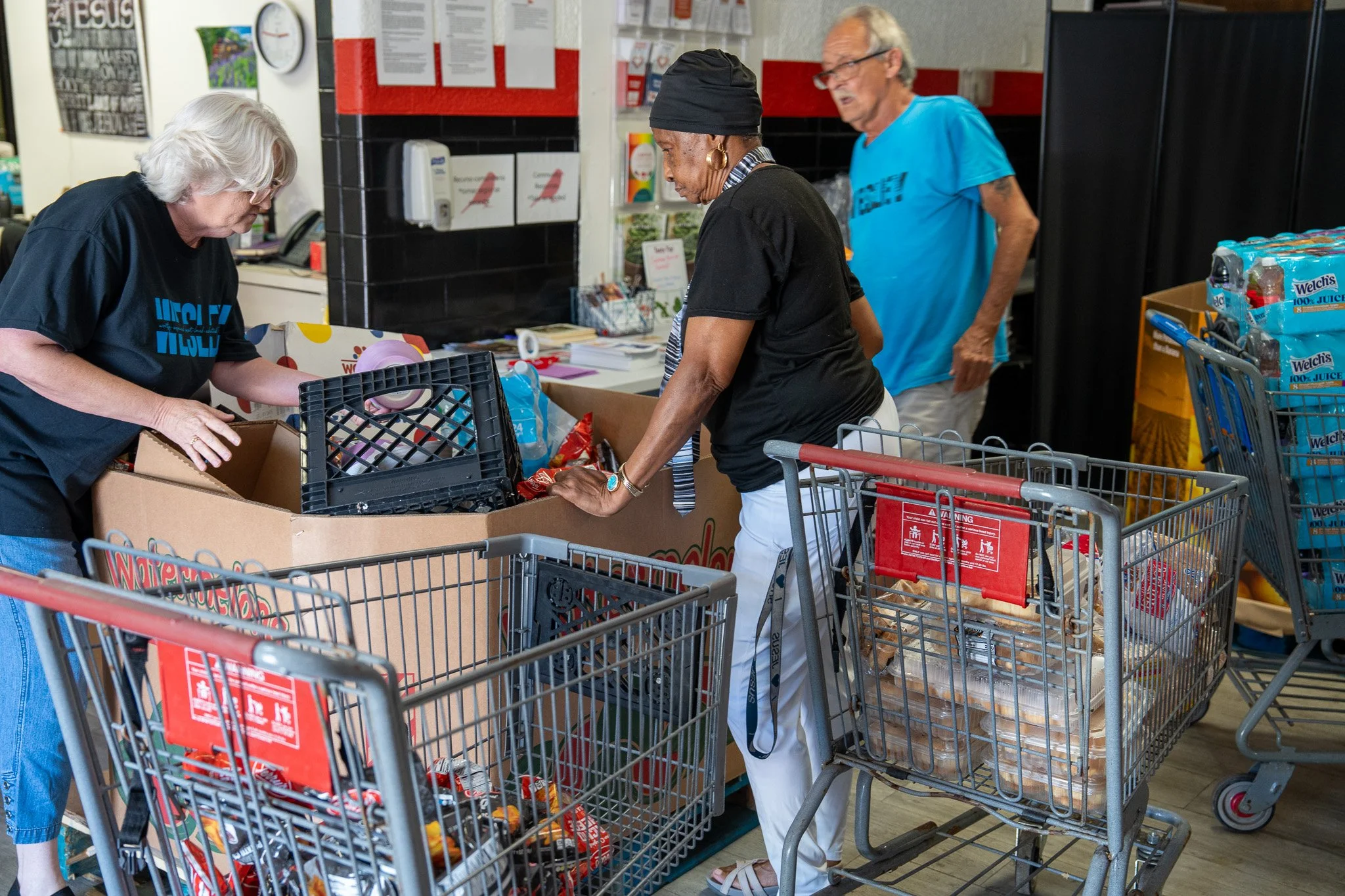 Three people are shopping at a grocery store checkout. They are loading items into shopping carts and a large cardboard box. One woman with white hair is on the left, a woman with dark hair and a head covering is in the middle, and a man with glasses
