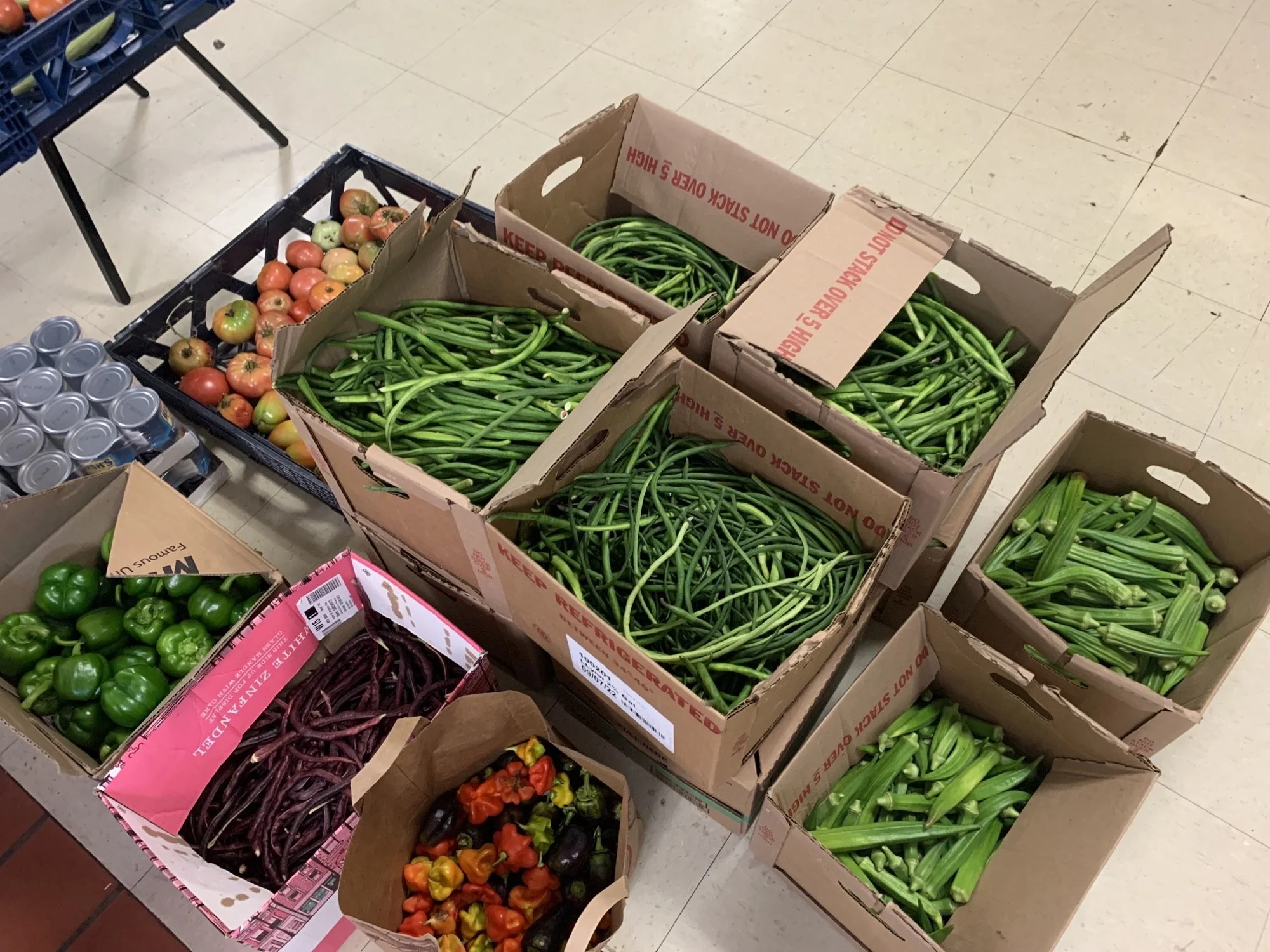 Boxes of fresh vegetables including green beans, okra, green bell peppers, and cherry tomatoes, along with cans of drinks on a light-colored tiled floor.