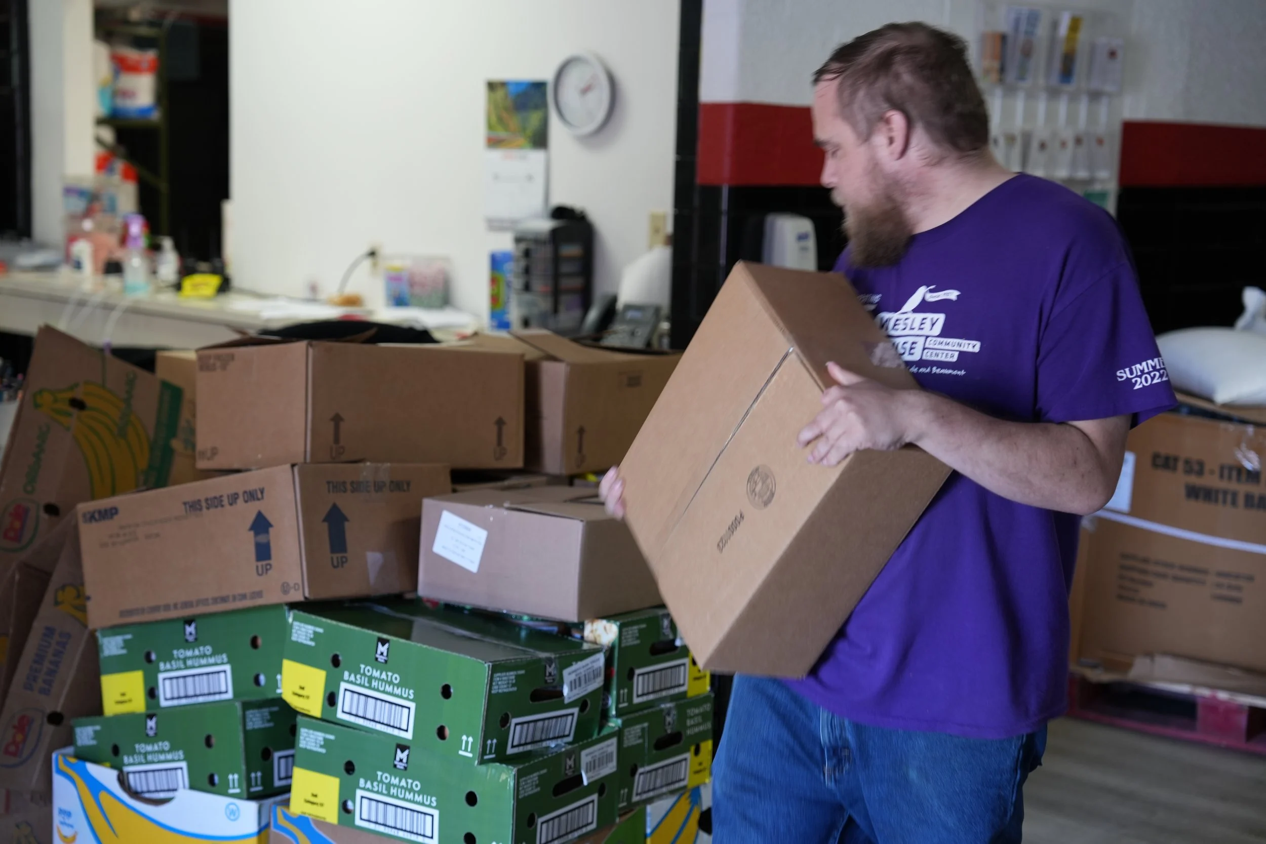 A man with a beard and short hair wearing a purple shirt and blue jeans is stacking cardboard boxes in a room filled with boxes, some labeled with vegetables like tomatoes, and a counter with office supplies and a clock on the wall.