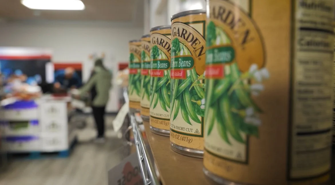 Cans of green beans displayed on a supermarket shelf, with shoppers in the background.