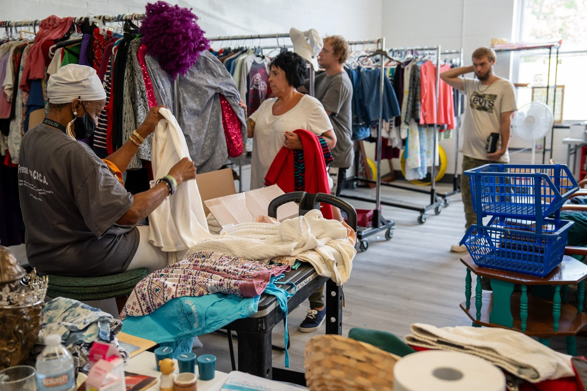 People shopping at a thrift store with clothing racks, tables with folded clothes, and shopping carts.