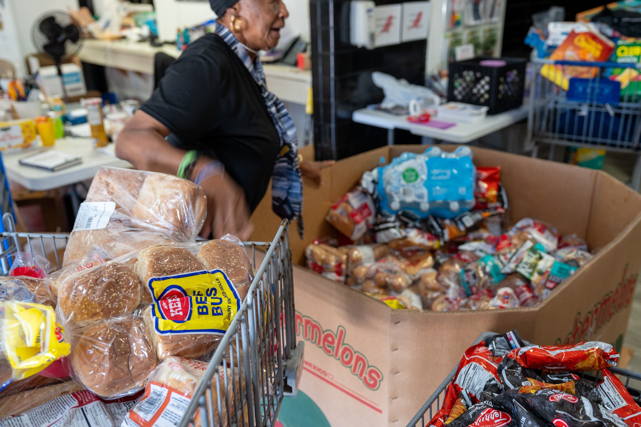 A woman shopping in a grocery store, with a cart filled with bread and snacks, and a large box containing packaged baked goods.
