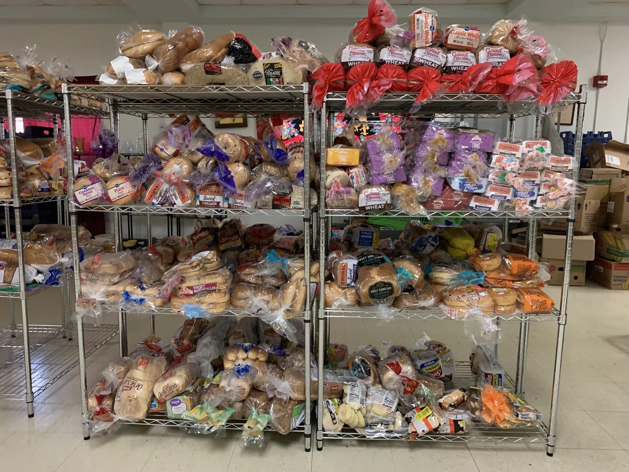 Metal shelving unit filled with various types of bread, including baguettes, hamburger buns, and sandwich bread, in plastic bags, in a storage area.