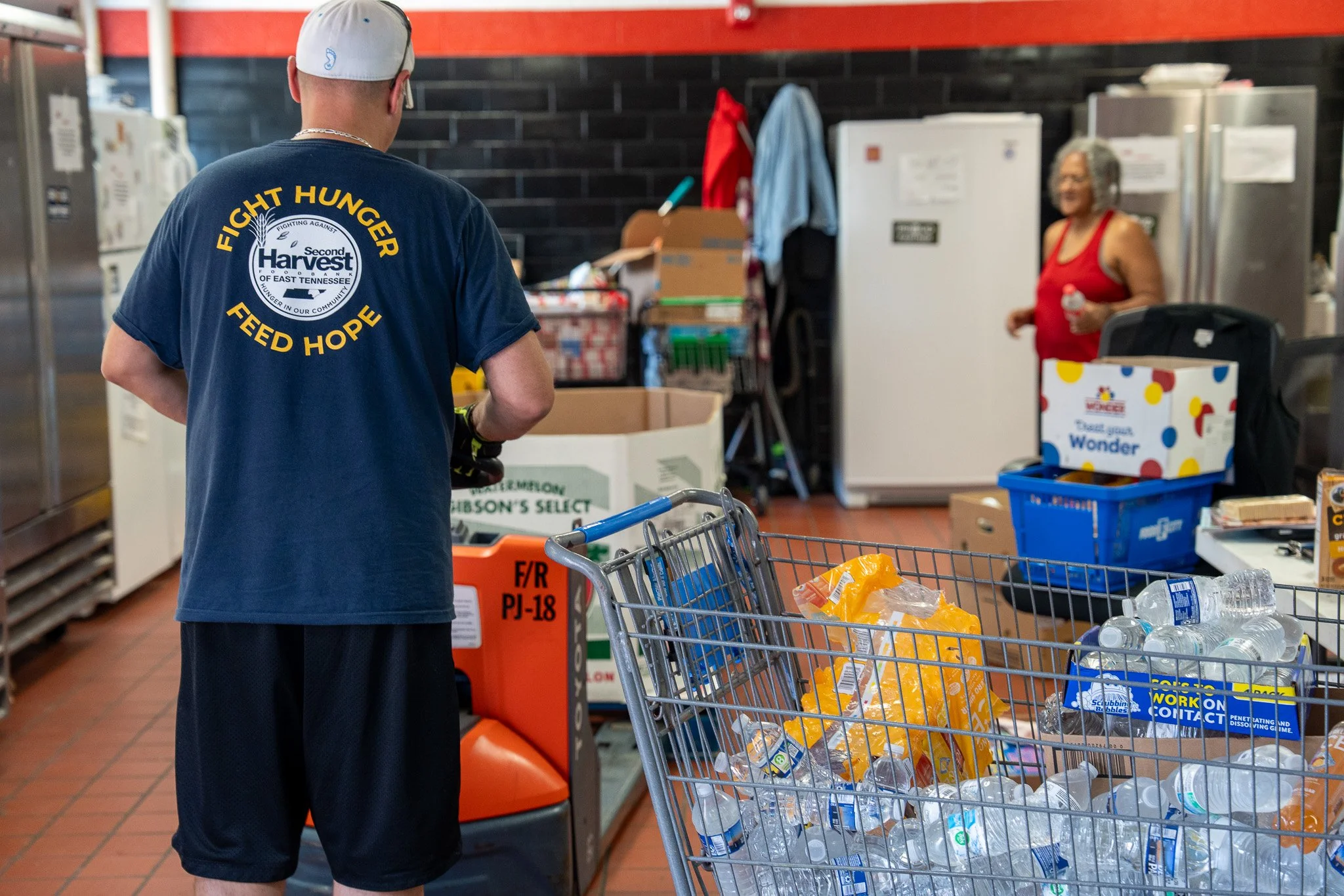 A young man wearing a navy blue T-shirt with yellow text and a white cap pushes a shopping cart filled with bottled water, food supplies, and other items in a grocery store or donation center. An older woman in a red tank top stands in the background