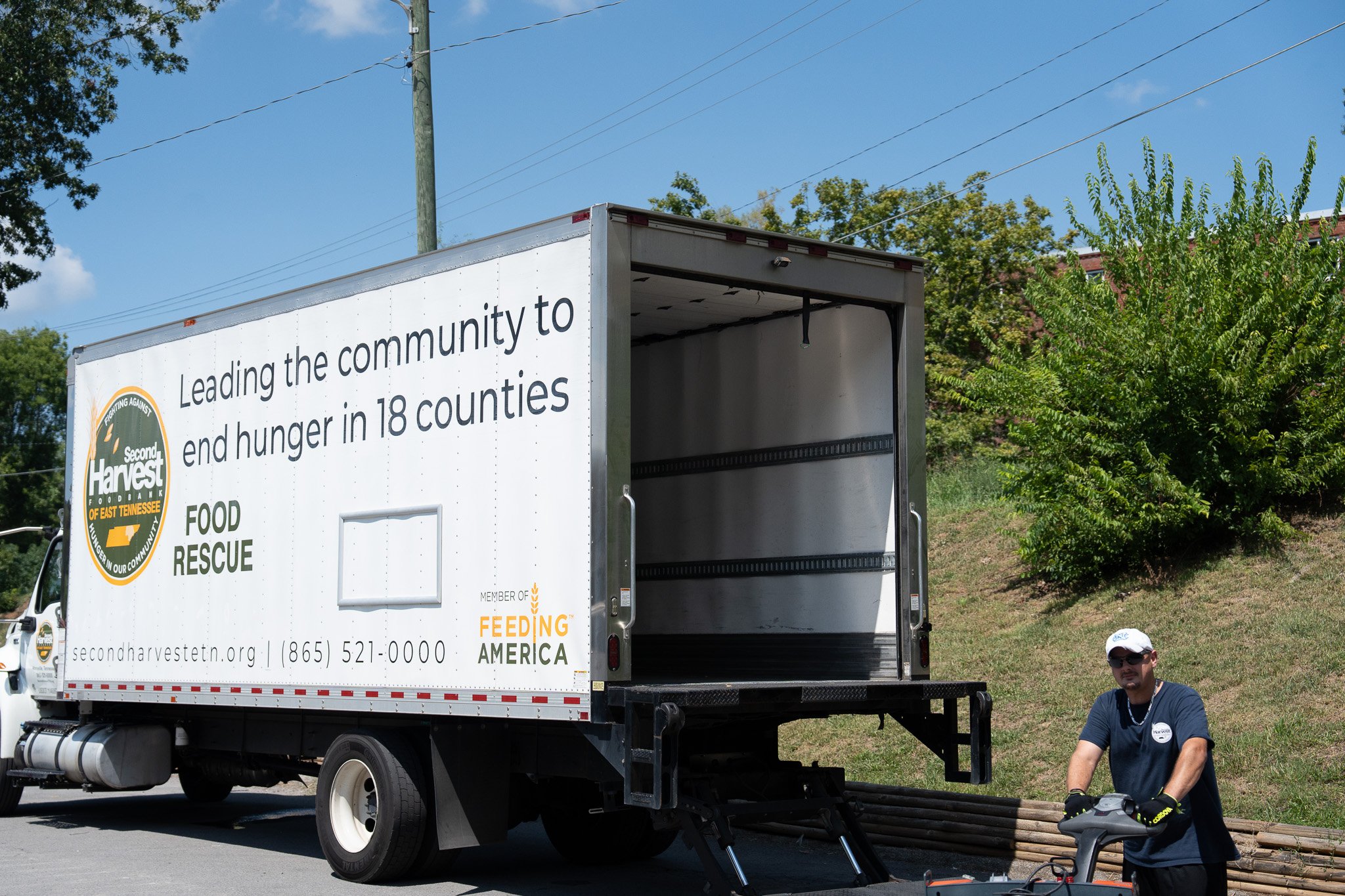 A food rescue truck with a logo reading 'Second Harvest Food Bank of East Tennessee' parked outdoors. A man wearing sunglasses, a white cap, and a blue shirt stands nearby with a bicycle, in front of a grassy hill with trees, under a clear blue sky.