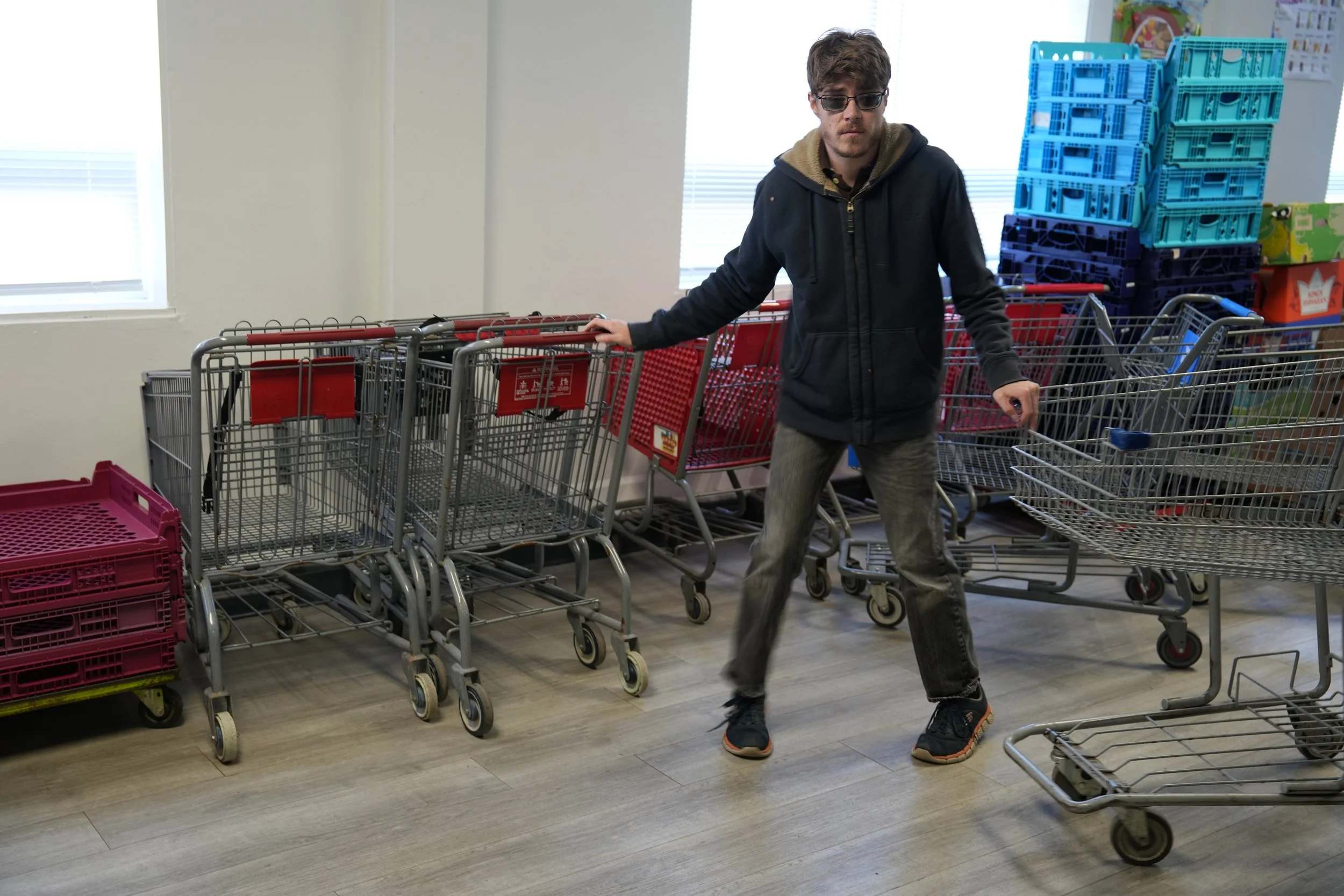 Young man with sunglasses wearing a black hoodie and jeans standing in a store aisle with shopping carts, some stacked with blue crates and others with pink crates, near a window with blinds.