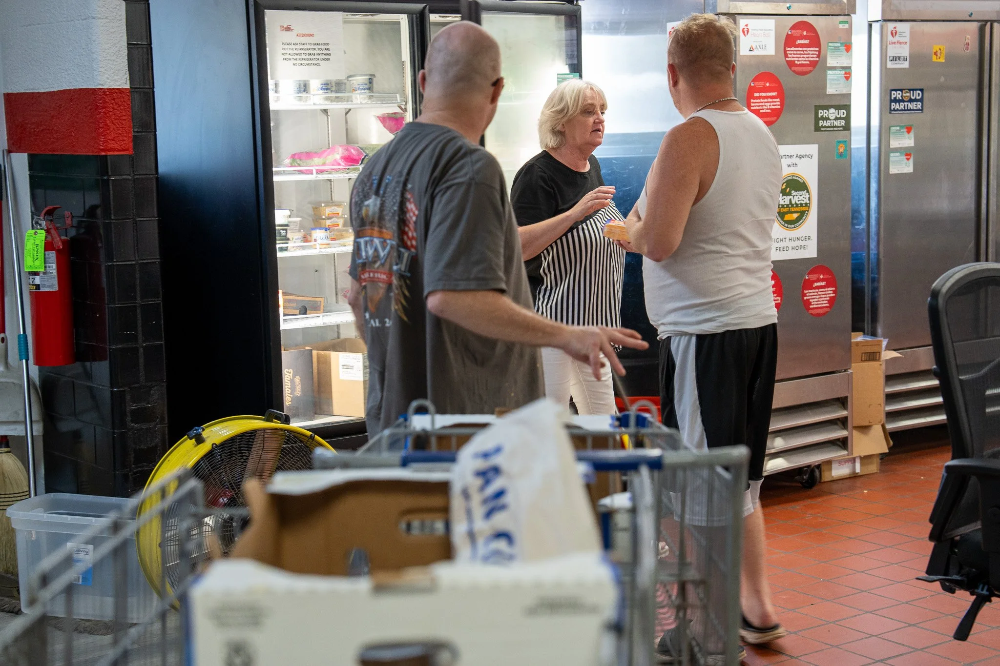 Three people engaged in conversation near a refrigerated display case in a store or food kitchen. One woman is speaking while two men listen. The woman has blonde hair and is wearing a black top with a striped apron. The men are casually dressed, one