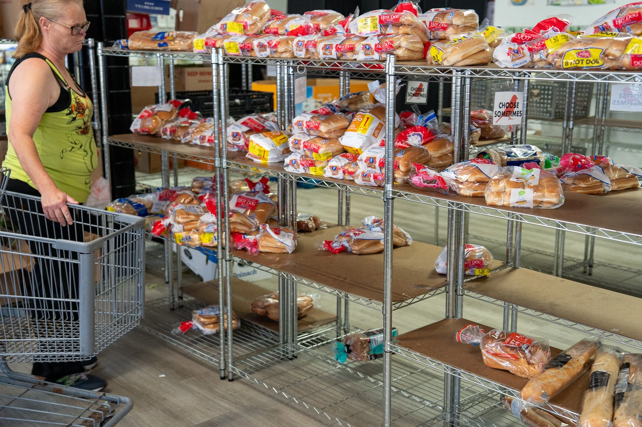 Empty bakery display shelves with loaves of bread and hot dog buns, and a woman with glasses and a yellow tank top pushing a shopping cart in a grocery store.