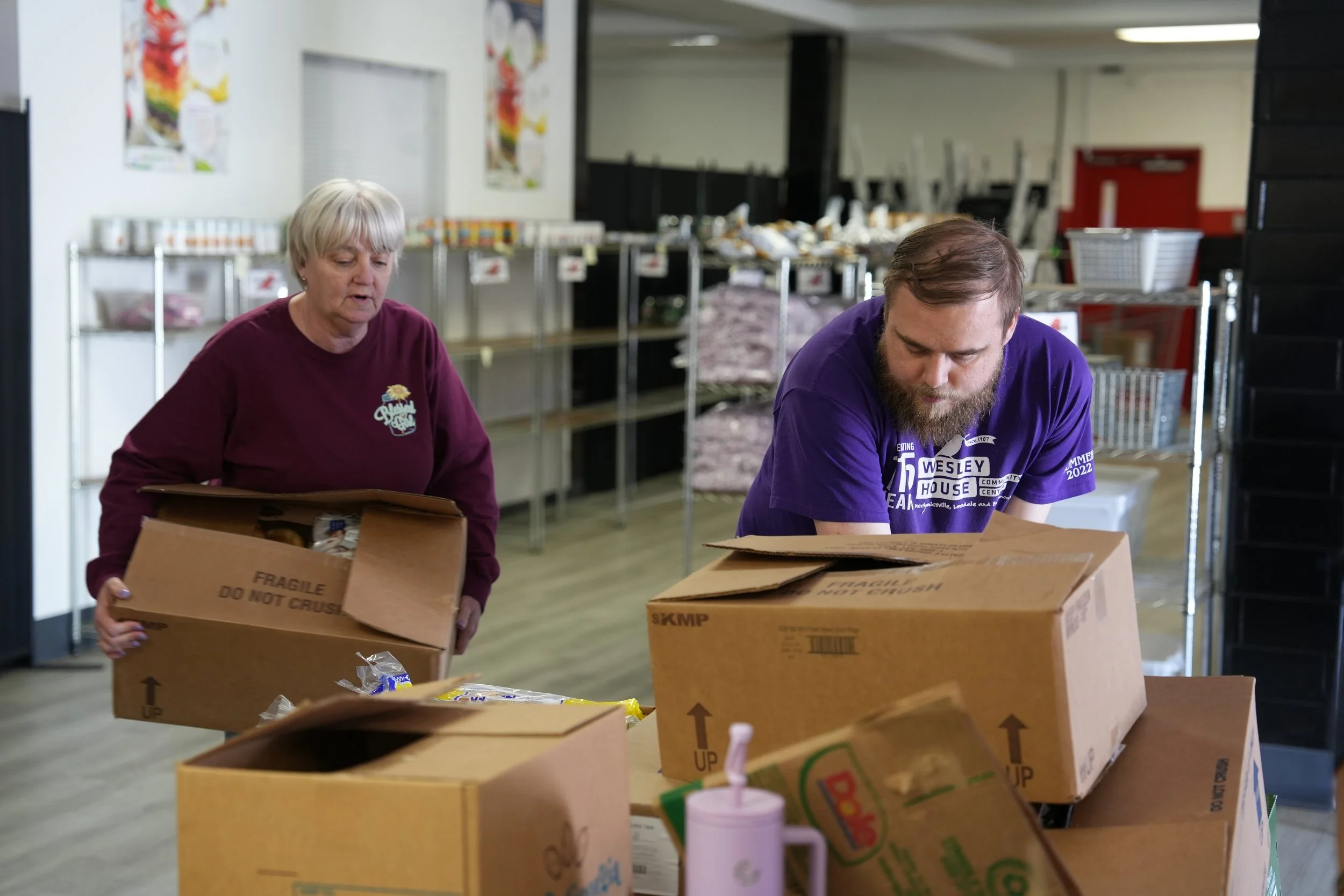 Two people packing boxes in a community center or food bank, with shelves and supplies in the background.