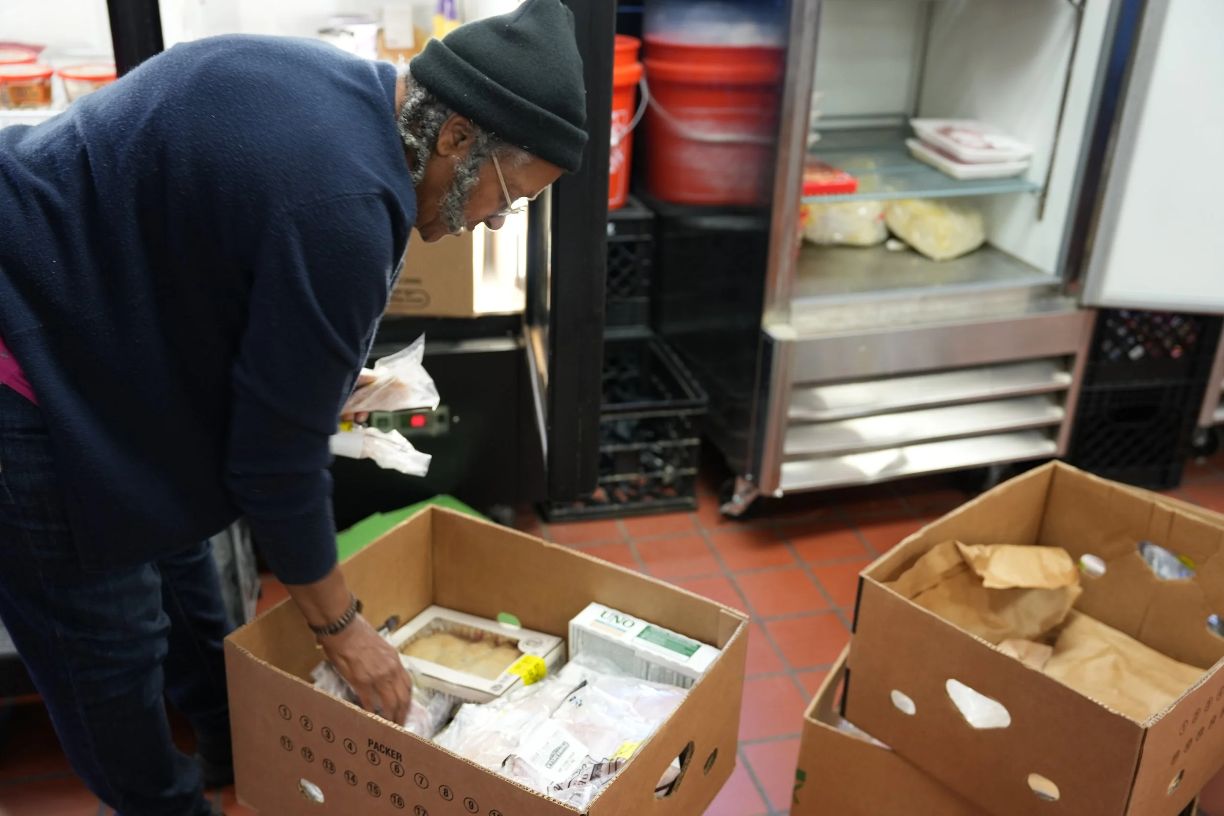 A man with gray hair, glasses, wearing a black beanie, a blue sweater, and dark plaid pants, is leaning over a large cardboard box filled with various packaged food items in a commercial kitchen or food storage room. There are shelves and a refrigera