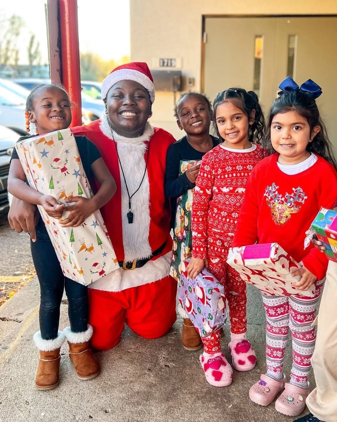A man dressed as Santa Claus kneeling on a sidewalk with four young girls in holiday pajamas, holding gifts and smiling outside a building.