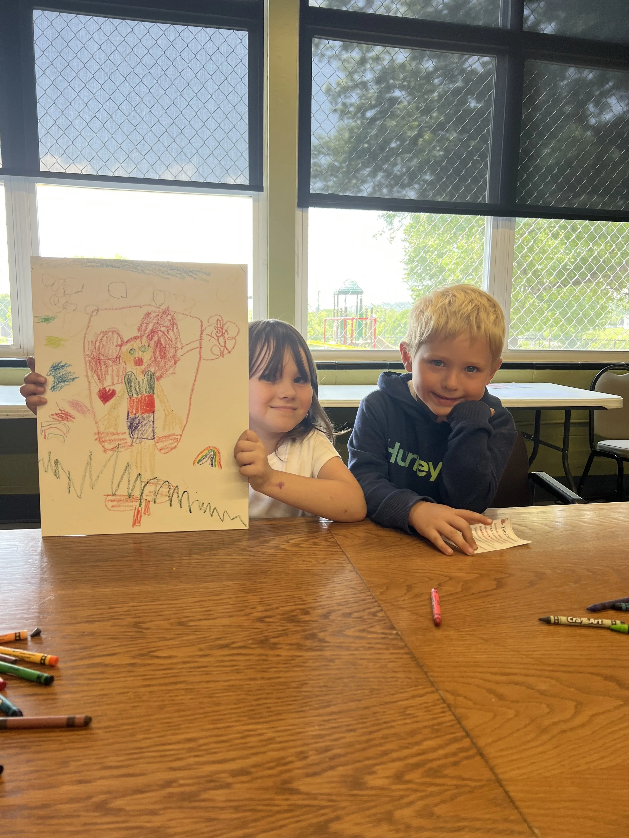Two children sitting at a wooden table with crayons, showing a colorful drawing of a girl with pigtails, hearts, a rainbow, and clouds.